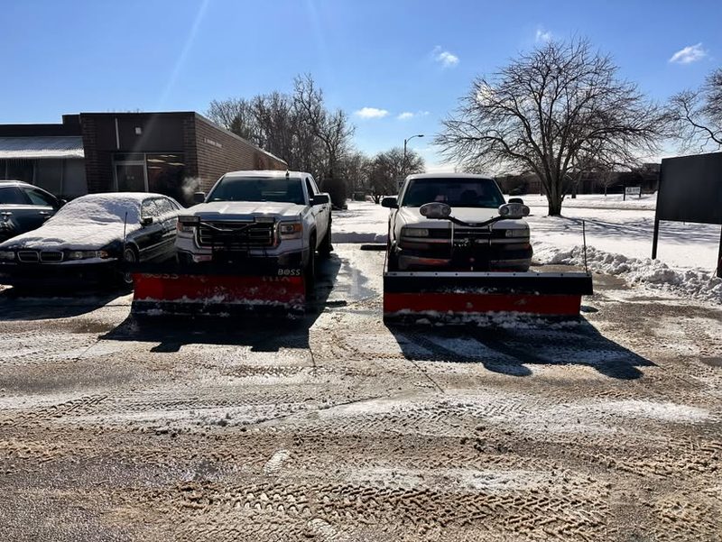 Two plow trucks with blades down on a cleared commercial lot.